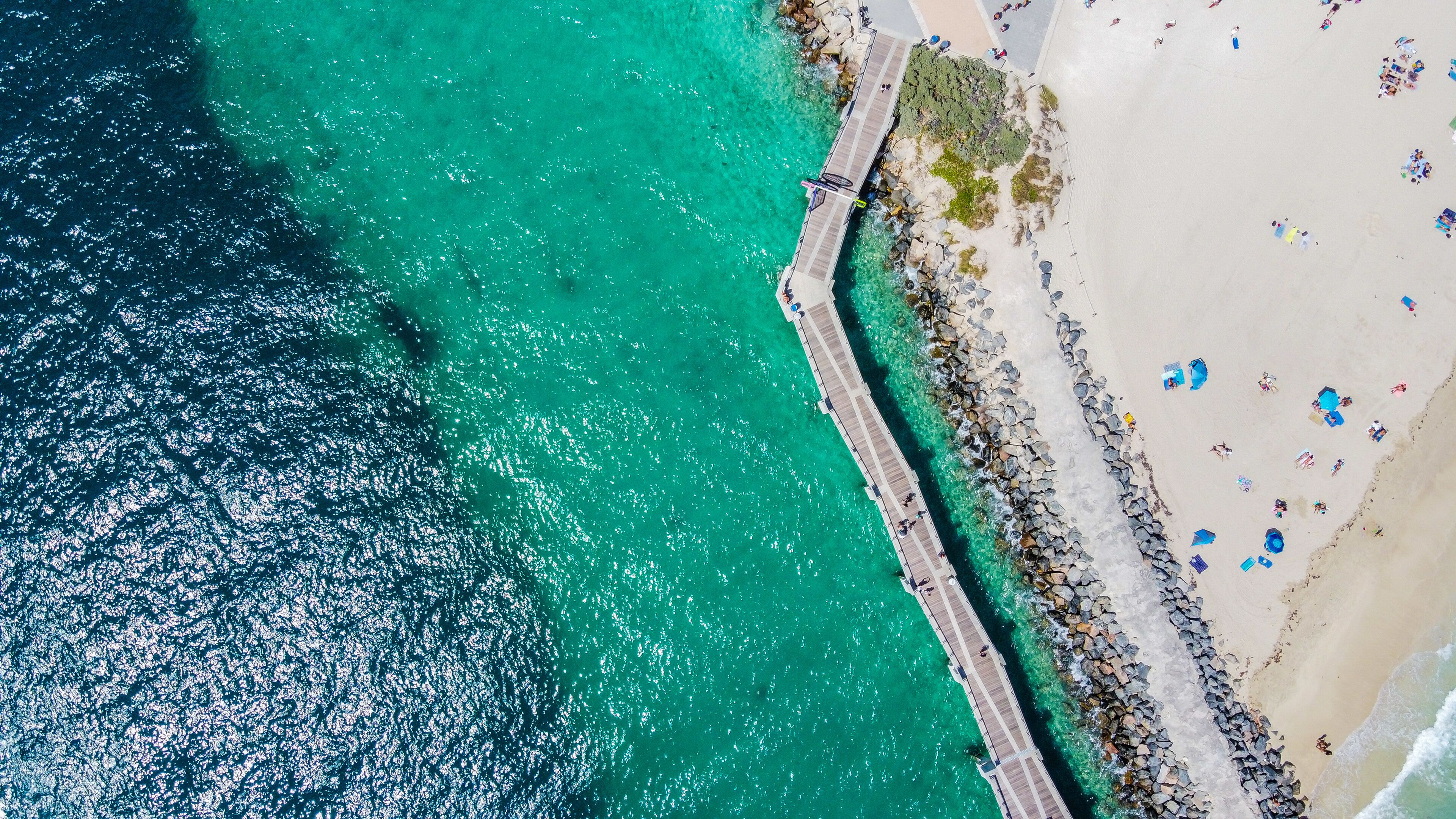 A coastal area with a pier extending into turquoise waters, sandy beach, and scattered umbrellas.