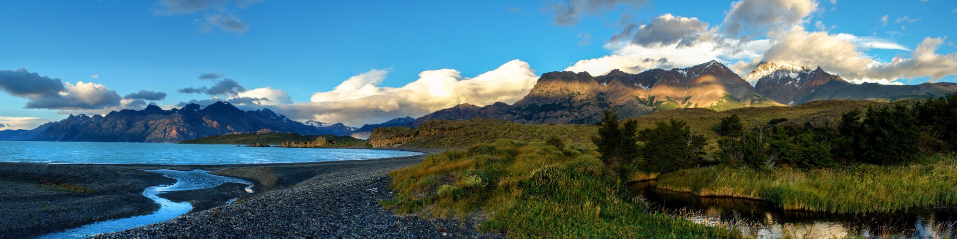 A coastal landscape with a pebbly beach, a calm sea, and a mountain range with snow-capped peaks under a partly cloudy sky.