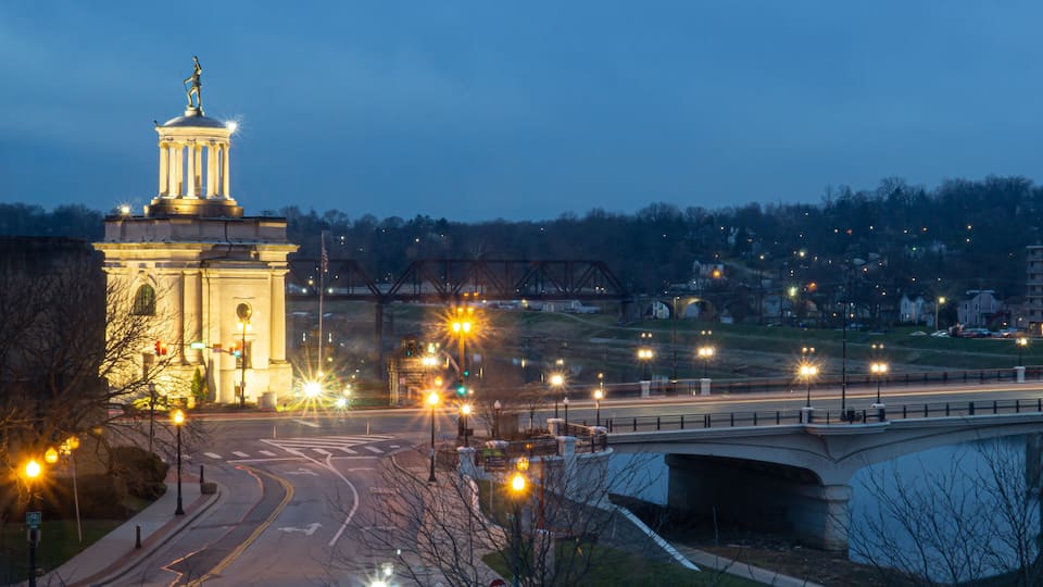 A cityscape at dusk with a prominent classical monument, a bridge, and illuminated streets.