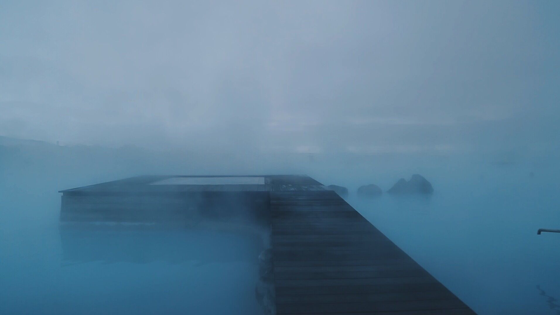 A wooden dock extending into a misty body of water with rocks visible in the background.