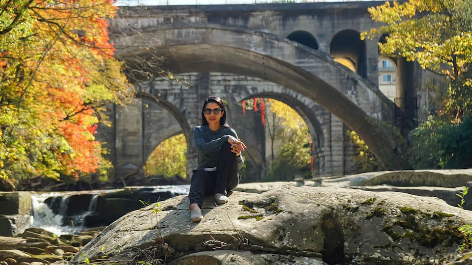 A person sitting on a rock by a river with a stone bridge in the background.
