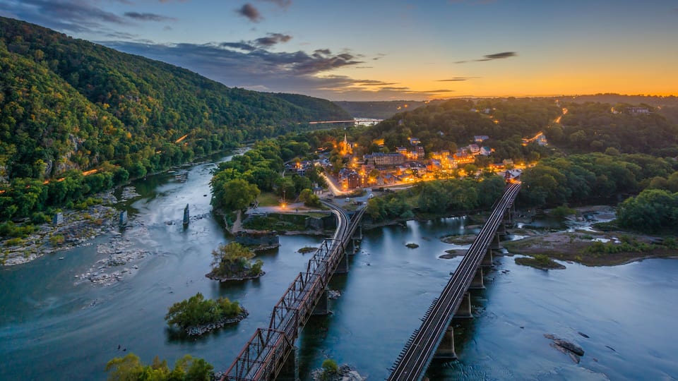 A river with a bridge, a town with illuminated buildings, and surrounding green hills.