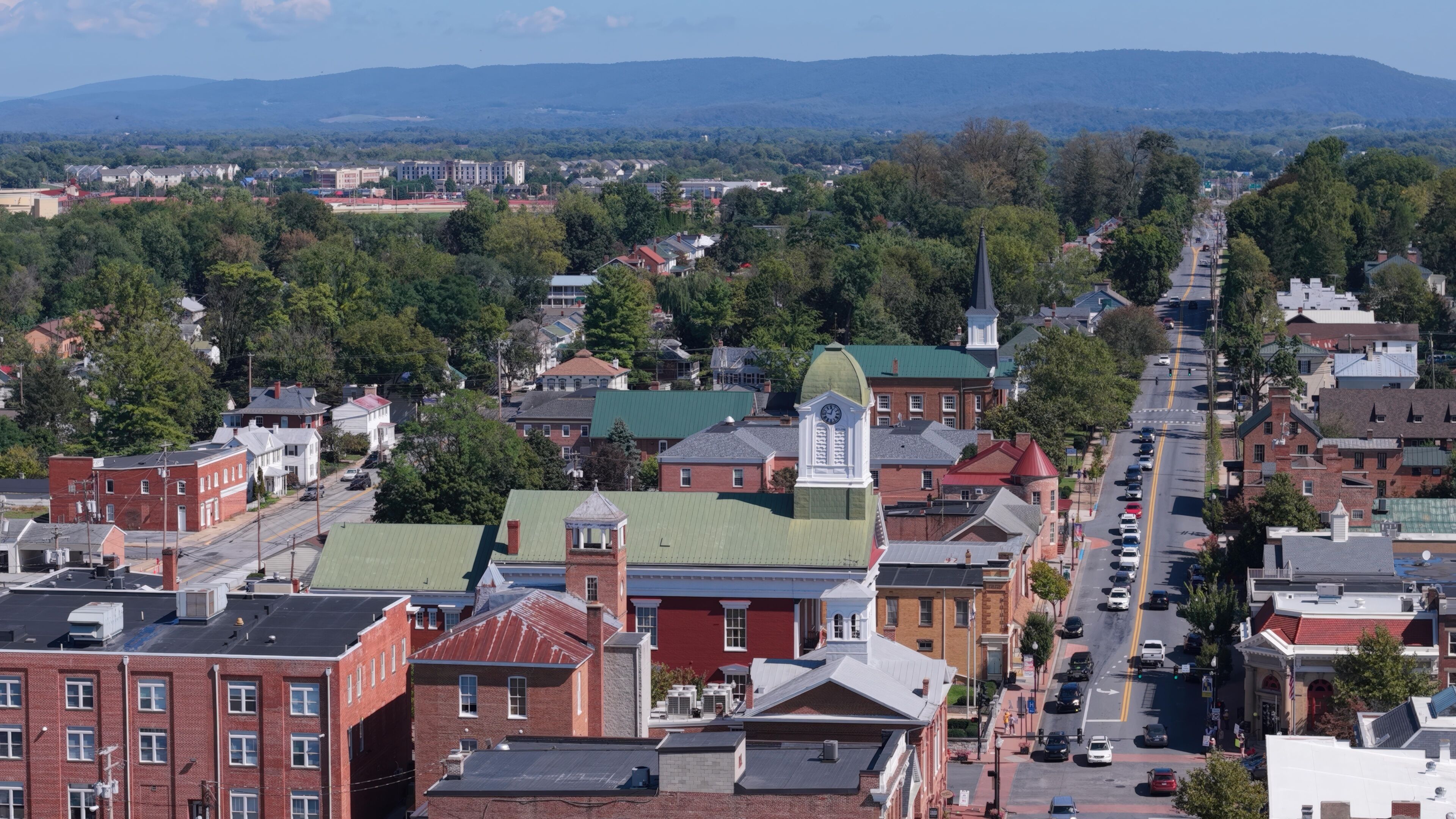 A town with a prominent clock tower, green-roofed buildings, and a mountainous backdrop.
