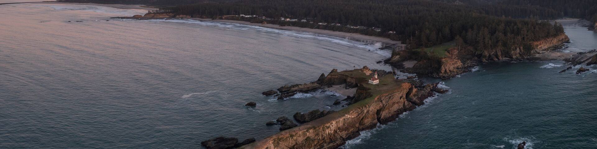 A coastal landscape with a lighthouse, rocky shoreline, and forested hills during sunset.