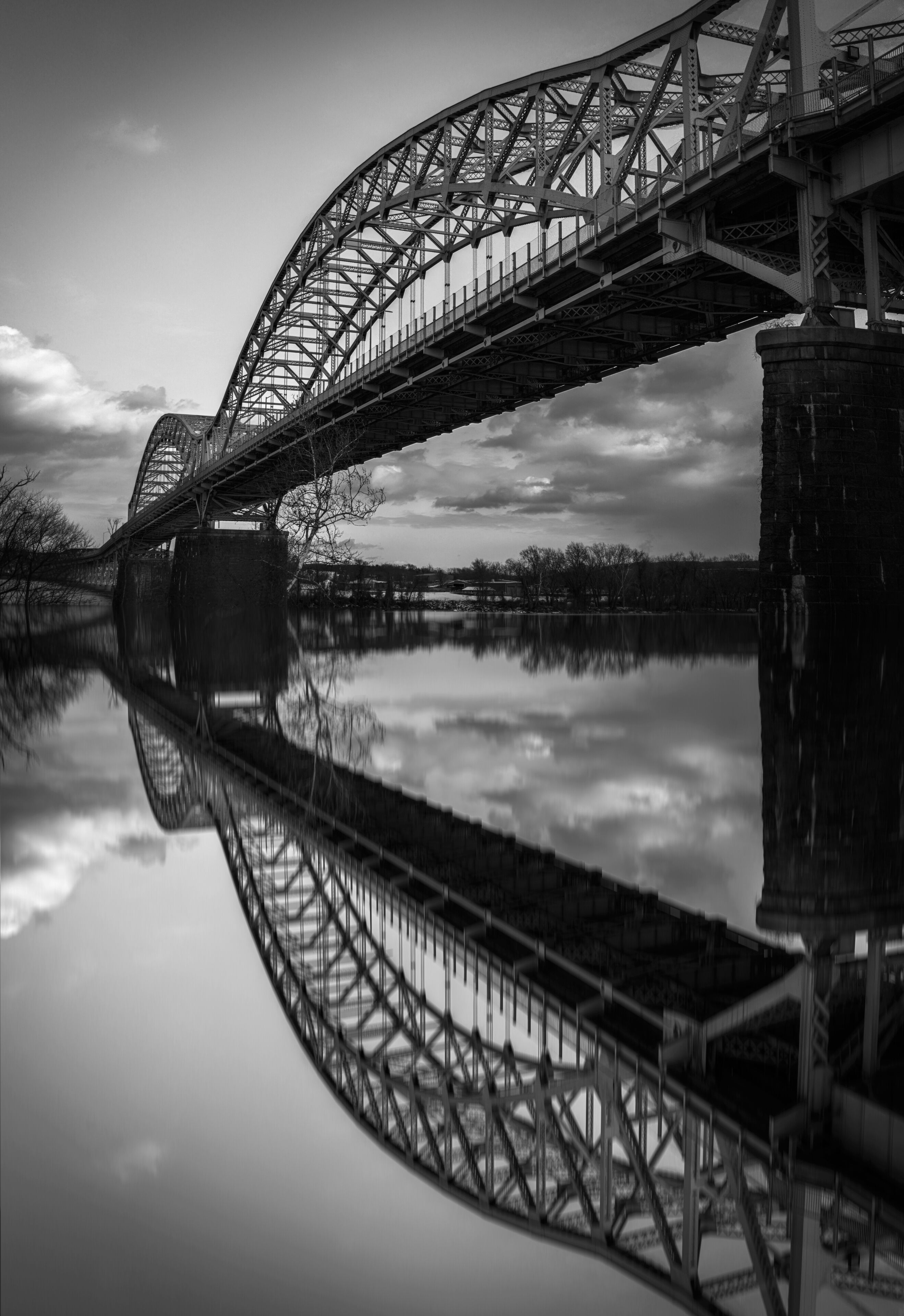 A large steel bridge spanning a calm river, with its reflection visible in the water.