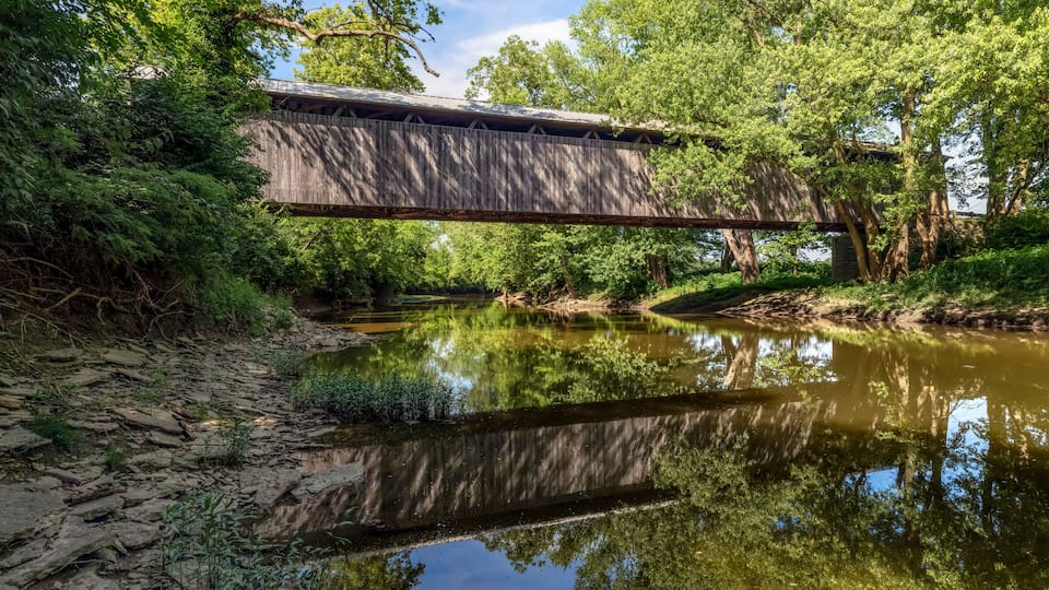 A wooden covered bridge over a calm river, surrounded by lush greenery and trees.