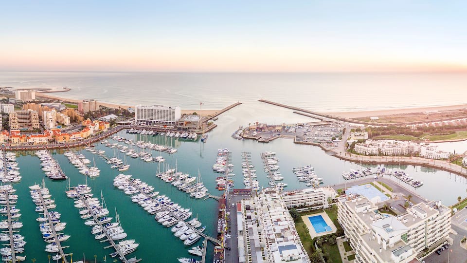 Un puerto deportivo lleno de barcos, un paisaje urbano costero y una puesta de sol sobre el océano.