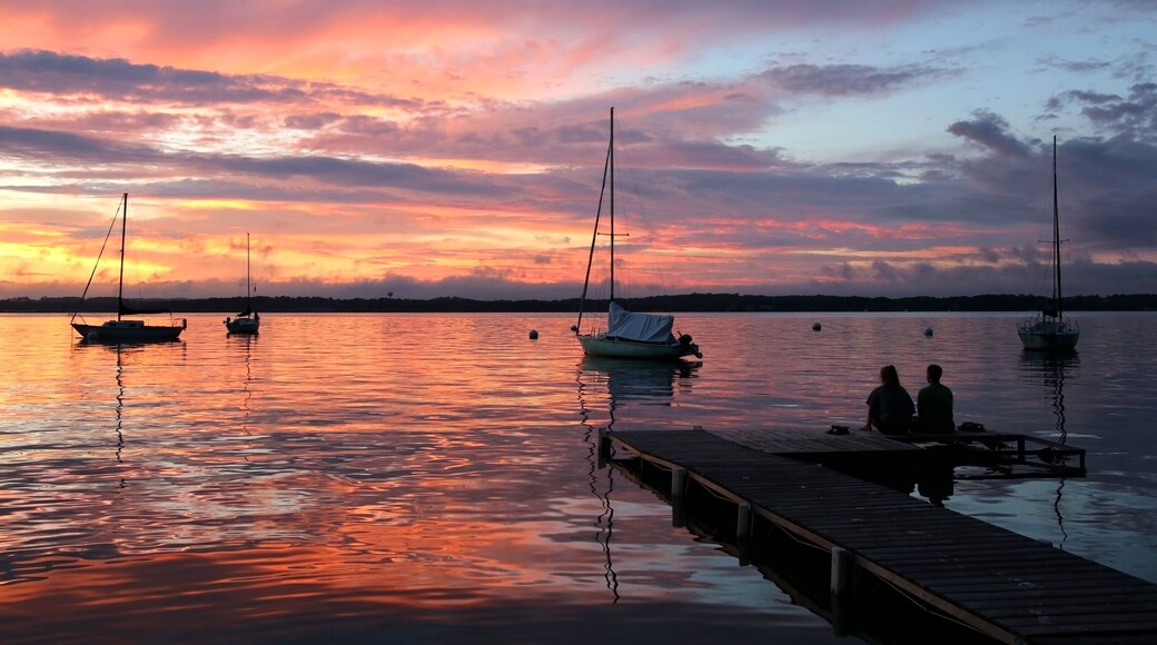 Amazing summer evening landscape with group of drifting yachts on a lake, two people on a wooden pier. Mendota during spectacular sunset. Bright sky reflects in the lake water.