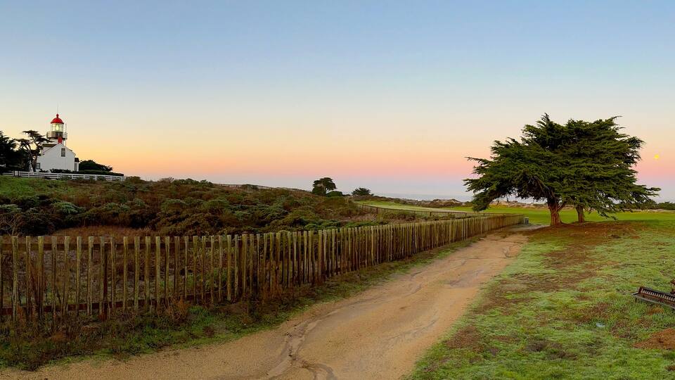 A coastal path with a lighthouse, wooden fence, and trees.