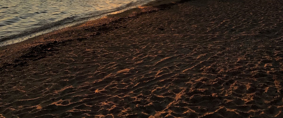 A sunset over a beach with rocks in the foreground and a sailboat in the distance.