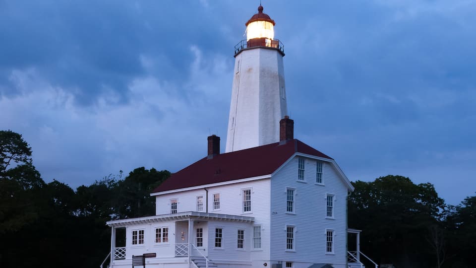A white lighthouse with a red roof, a small adjacent building, and a grassy area with trees in the background.