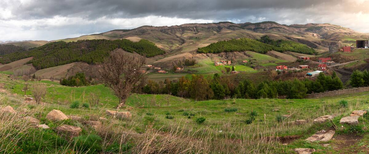 A mountainous landscape with rolling hills, scattered trees, and a few buildings in the valley.