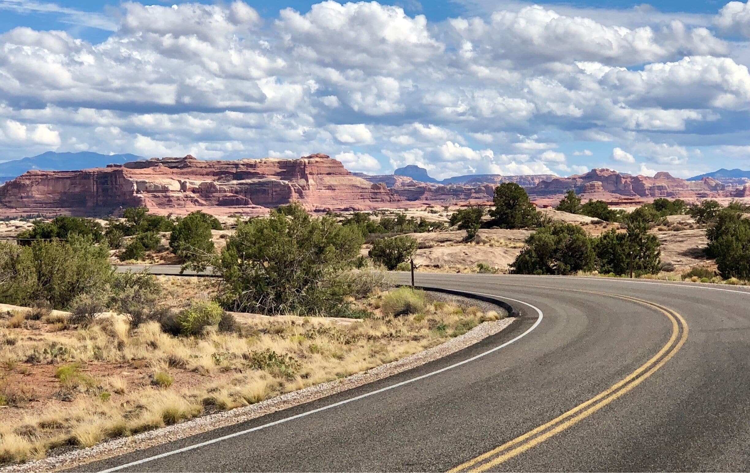 A winding road through a desert landscape with red rock formations and sparse vegetation under a partly cloudy sky.