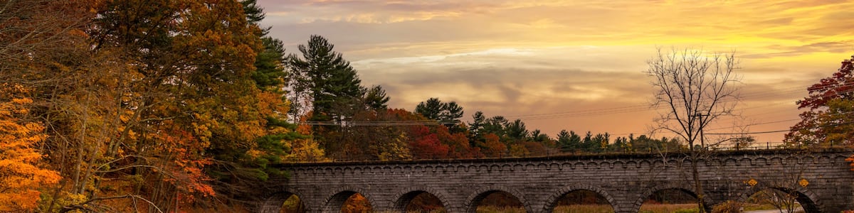 A stone bridge over a calm river with autumn foliage and a sunset sky.