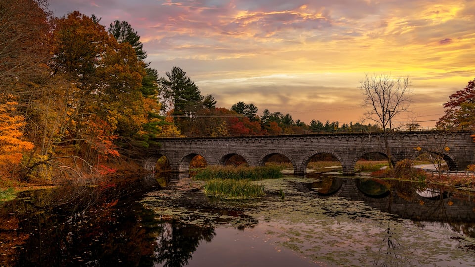 A stone bridge over a calm river with autumn foliage and a sunset sky.