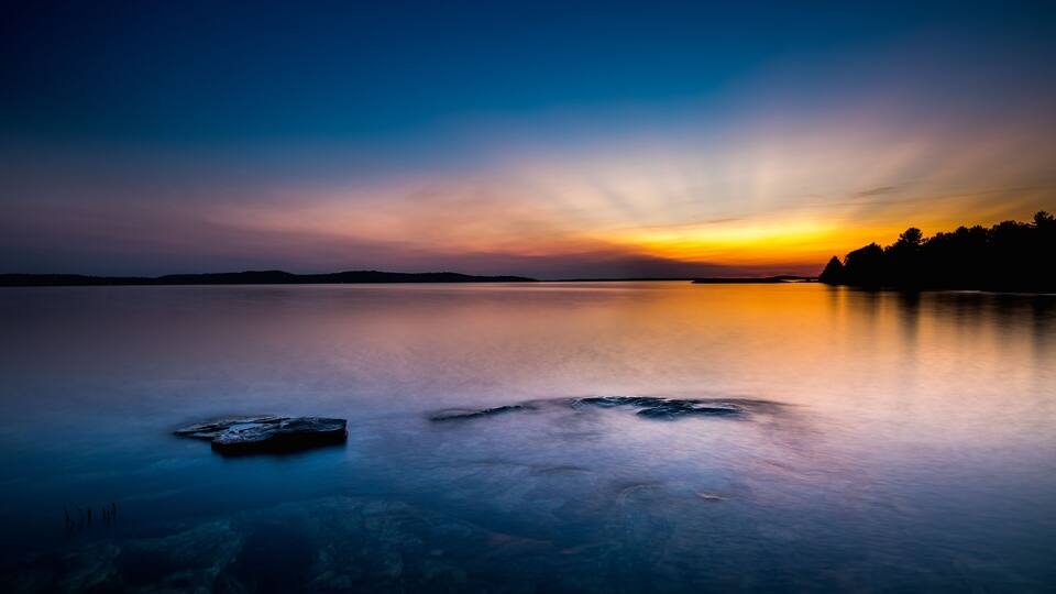 A calm lake at sunset with a silhouette of trees on the horizon.