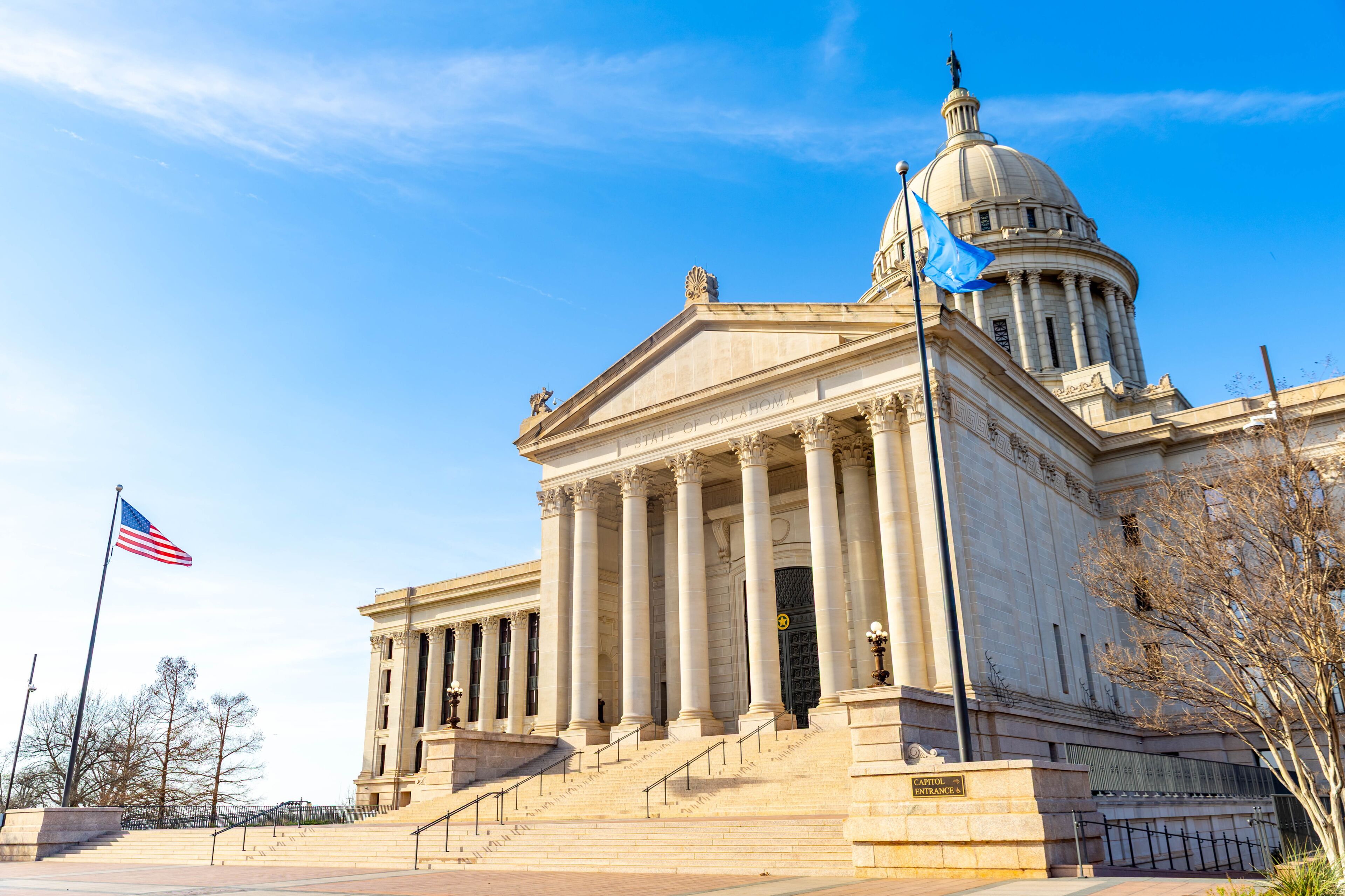 Un edificio neoclásico con una cúpula y columnas, una bandera estadounidense y un letrero que dice 'Oklahoma State Capitol'.