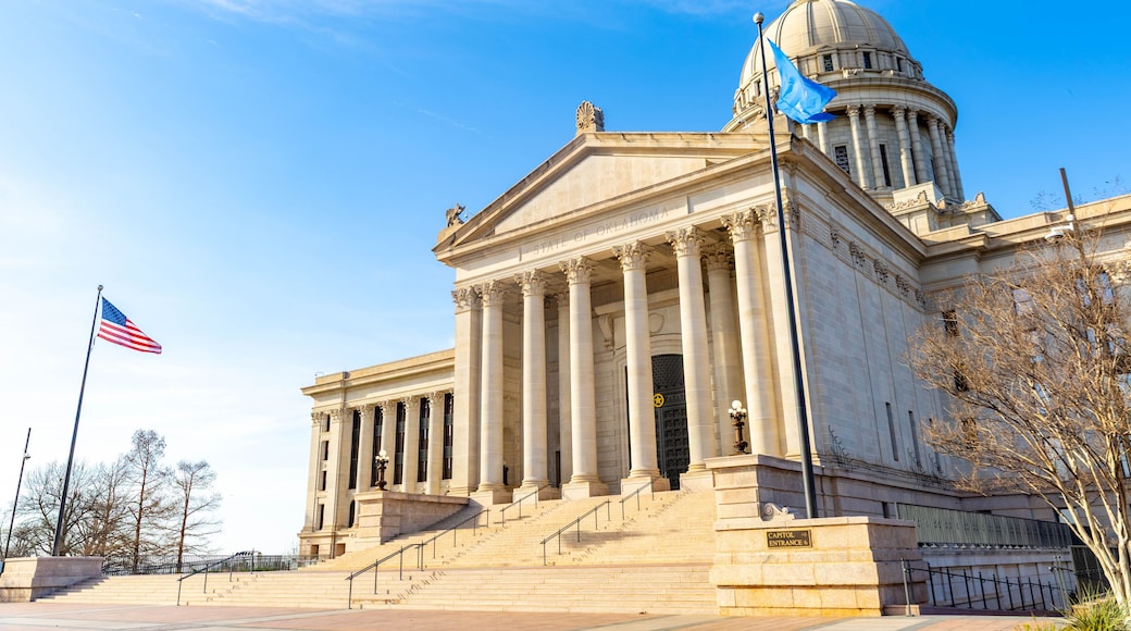 Un edificio neoclásico con una cúpula y columnas, una bandera estadounidense y un letrero que dice 'Oklahoma State Capitol'.