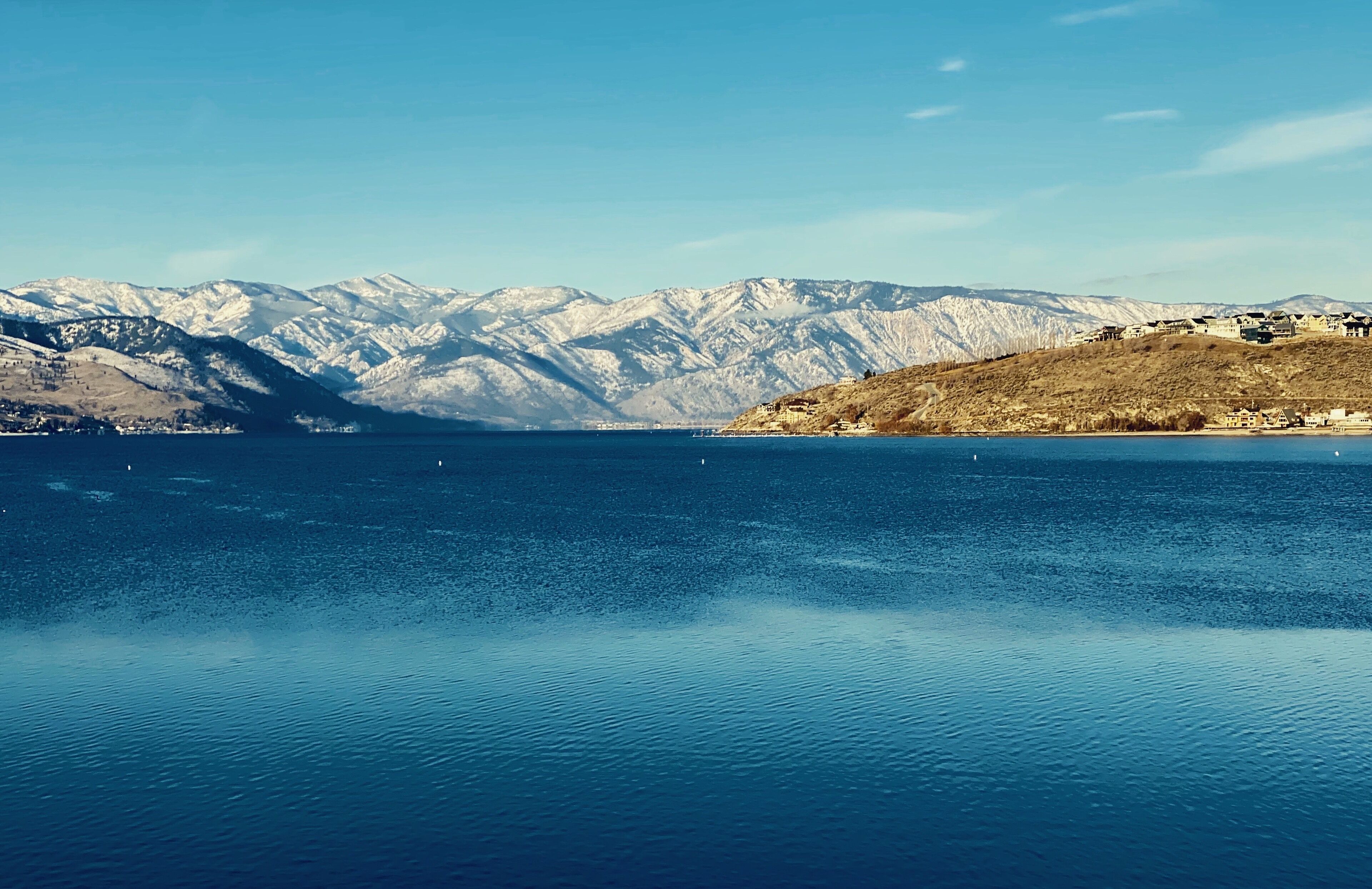 A calm lake with snow-capped mountains in the background and a few buildings on the right side.