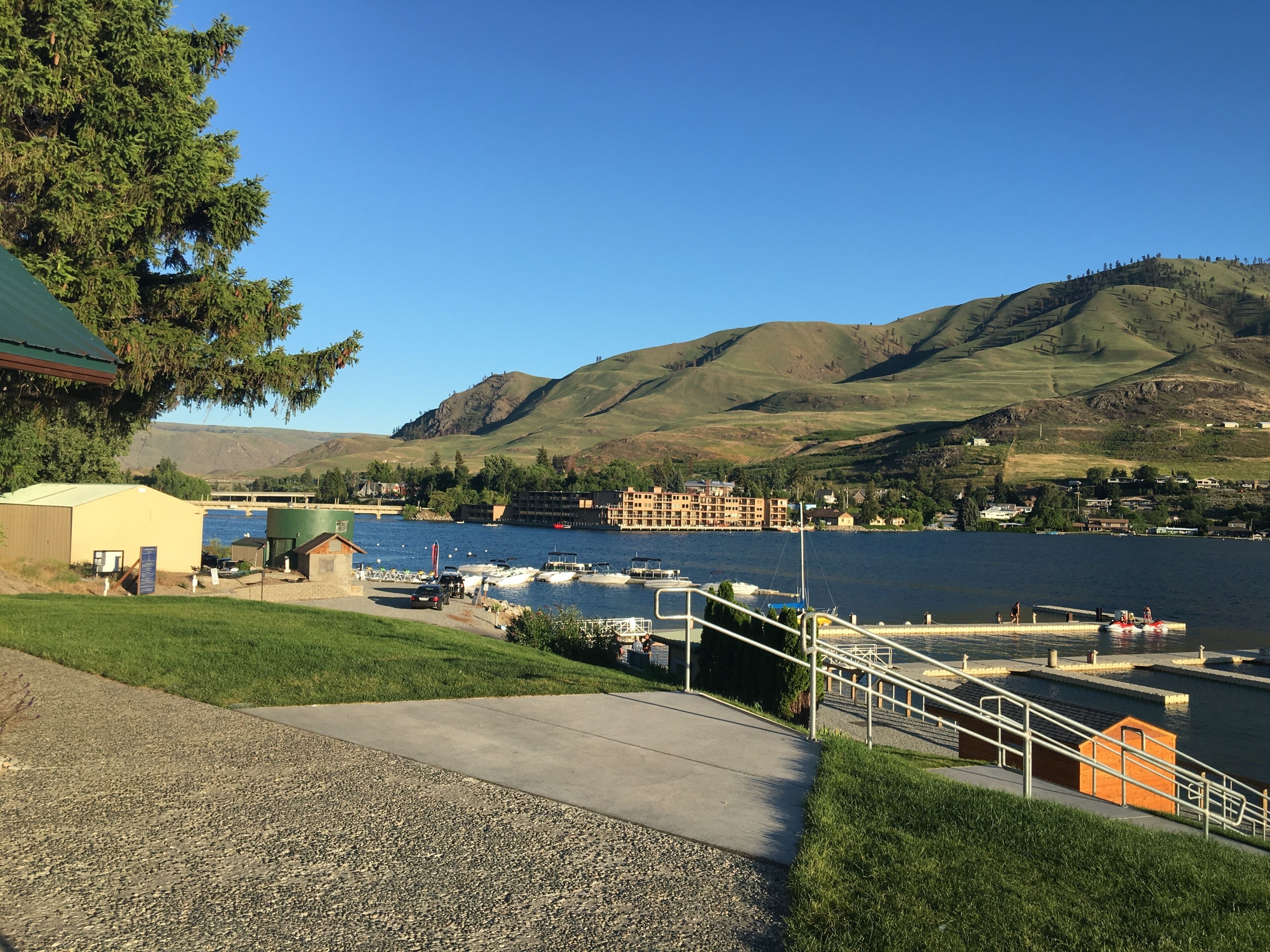 A lakeside view with a dock, boats, a small building, and hills in the background.