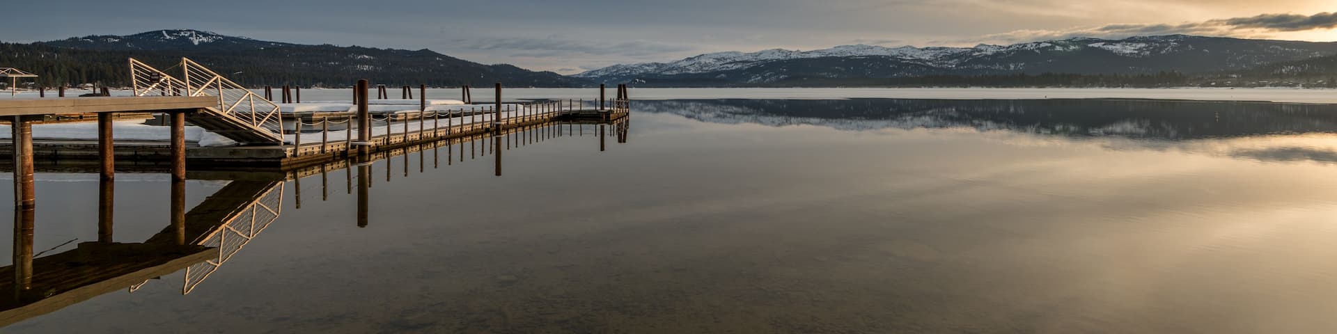 A calm lake with a wooden dock extending into the water, surrounded by snow-capped mountains and a clear sky.