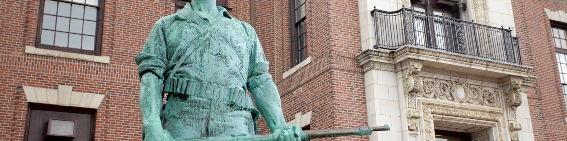 A statue of a man in uniform holding a rifle in front of a brick building with a balcony.