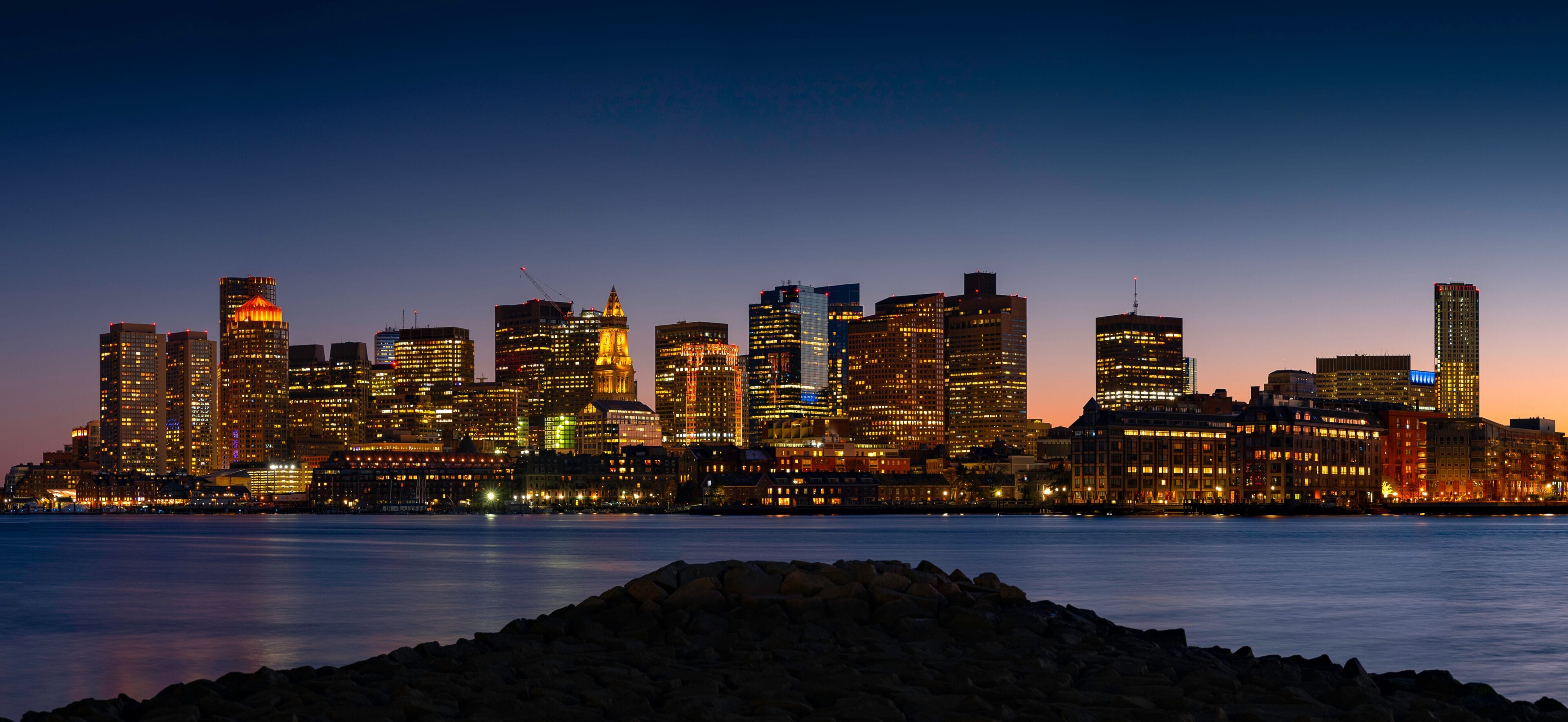 A city skyline at dusk with illuminated buildings along a waterfront.