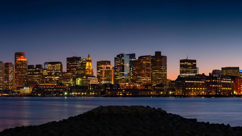A city skyline at dusk with illuminated buildings along a waterfront.