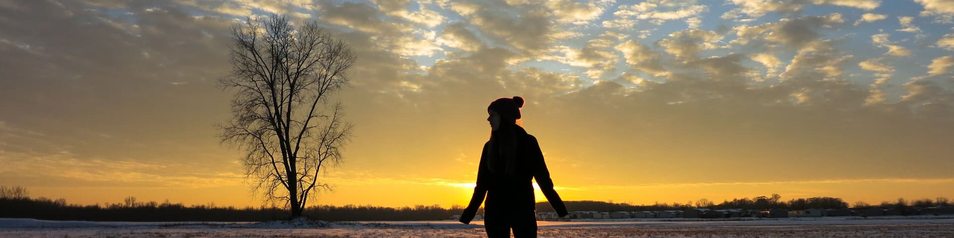 A person standing in a snowy field with a tree and a sunset in the background.