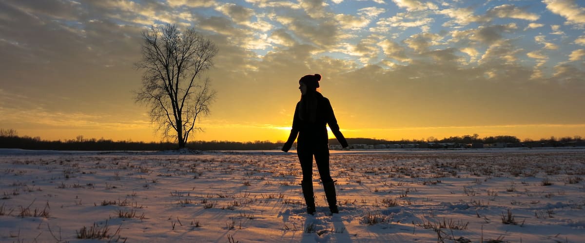 A person standing in a snowy field with a tree and a sunset in the background.