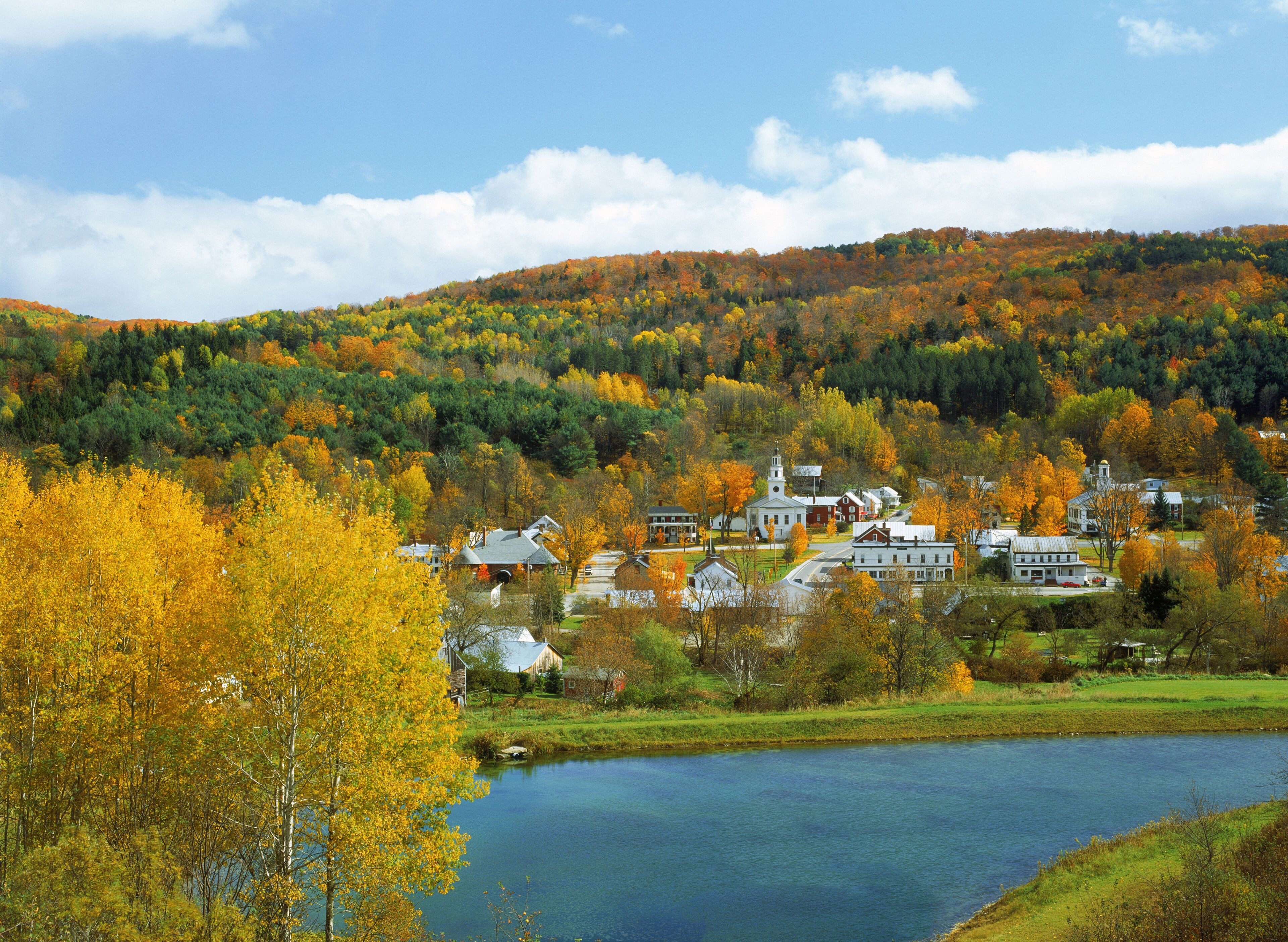 A small town with a church, houses, and a lake surrounded by autumn-colored trees.