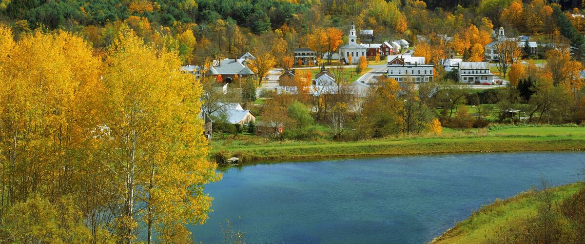 A small town with a church, houses, and a lake surrounded by autumn-colored trees.