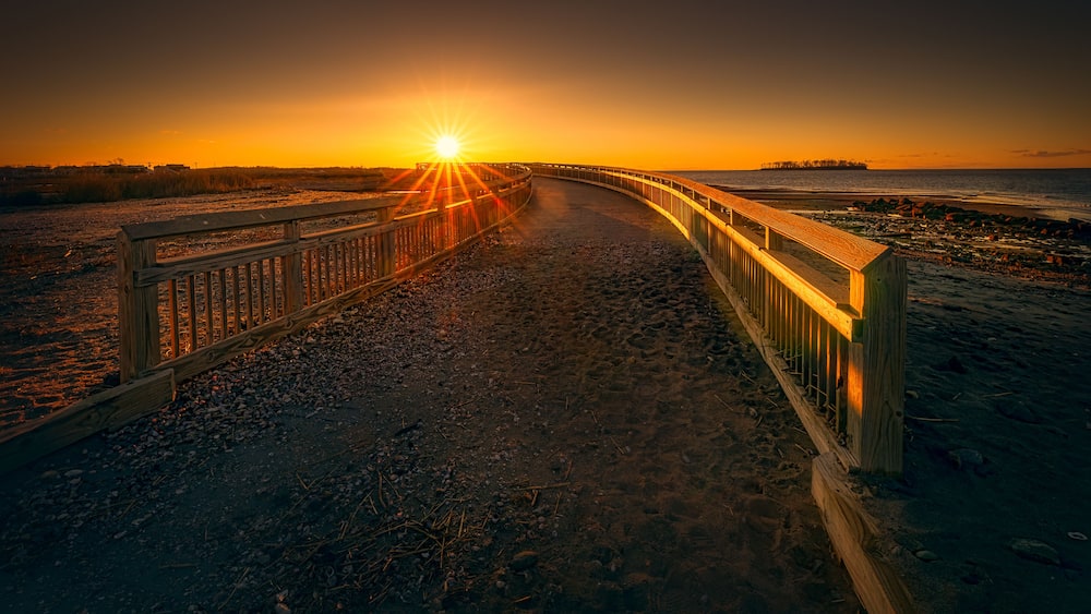 A wooden boardwalk leading to a sunset over a beach with a pier in the distance.