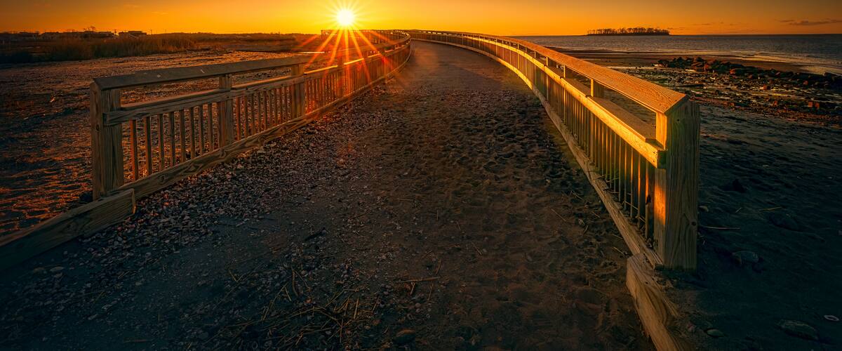 Ein hölzerner Steg führt zu einem Sonnenuntergang über einem Strand, in der Ferne ist ein Pier zu sehen.