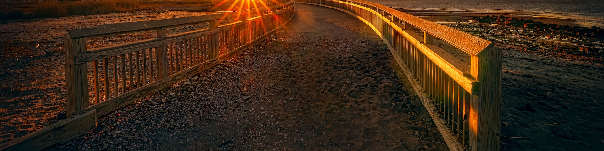 Una pasarela de madera que lleva hacia una puesta de sol sobre una playa, con un muelle a lo lejos.