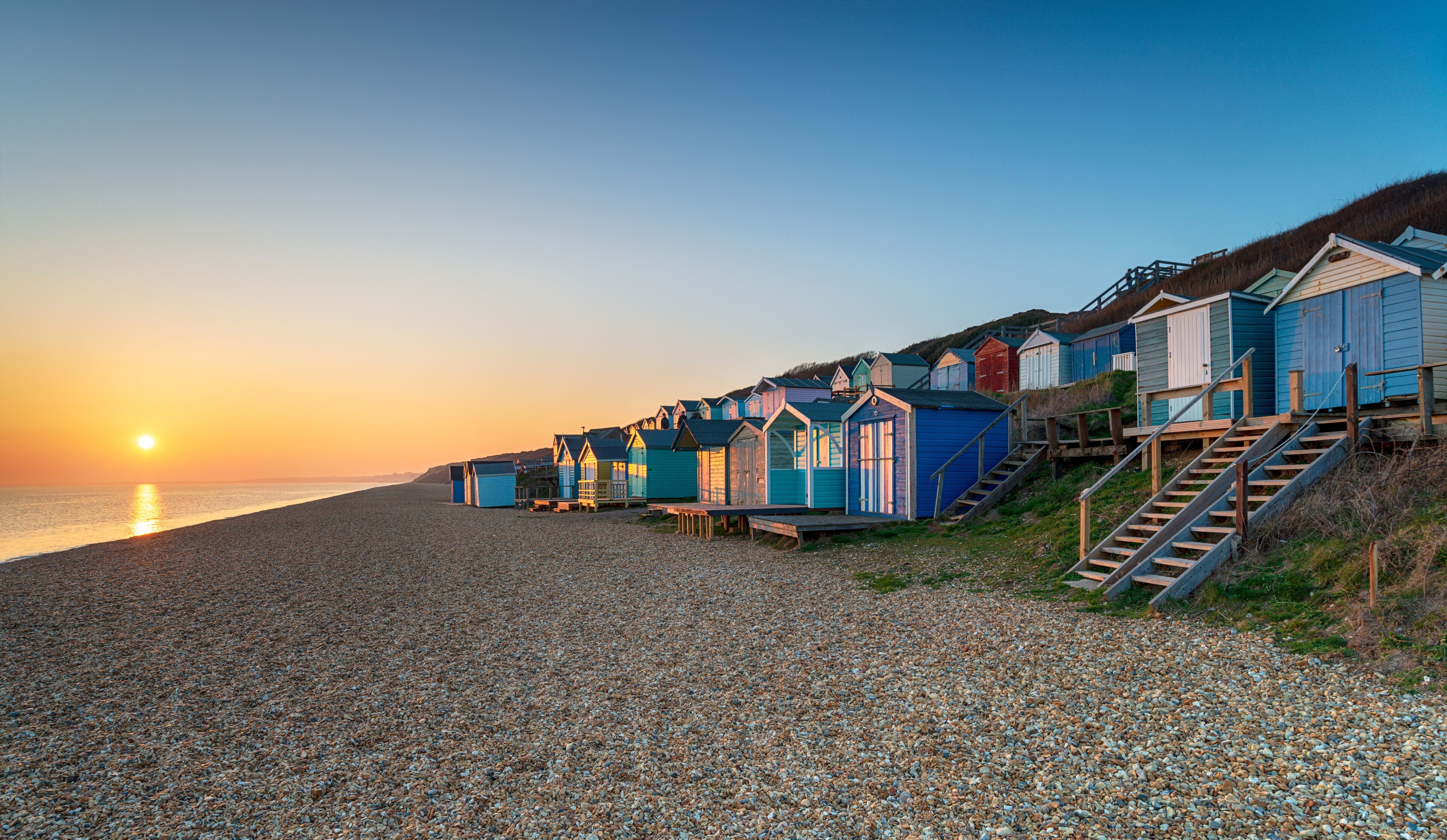 A pebbled beach with colorful beach huts and a sunset in the background.