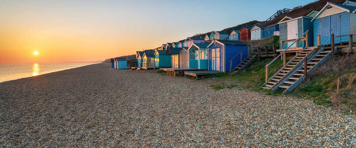 A pebbled beach with colorful beach huts and a sunset in the background.