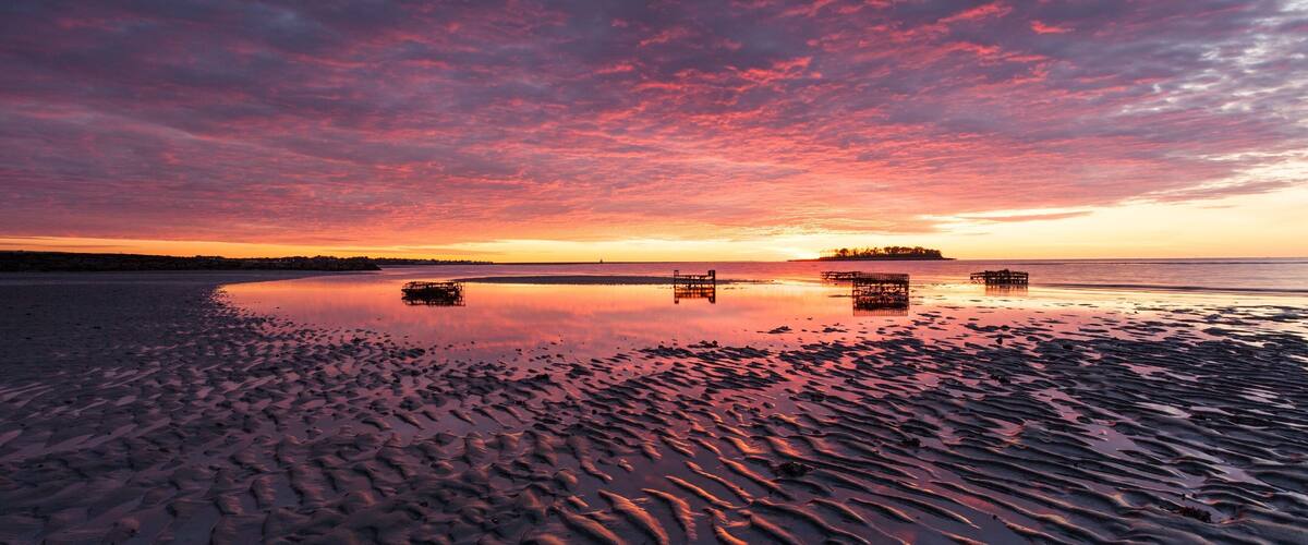 A sunset over a calm beach with rippled sand and distant boats.