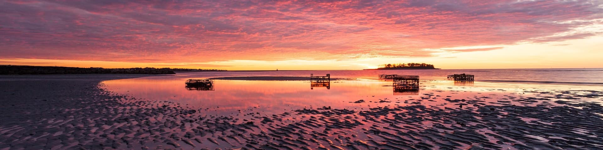 A sunset over a calm beach with rippled sand and distant boats.
