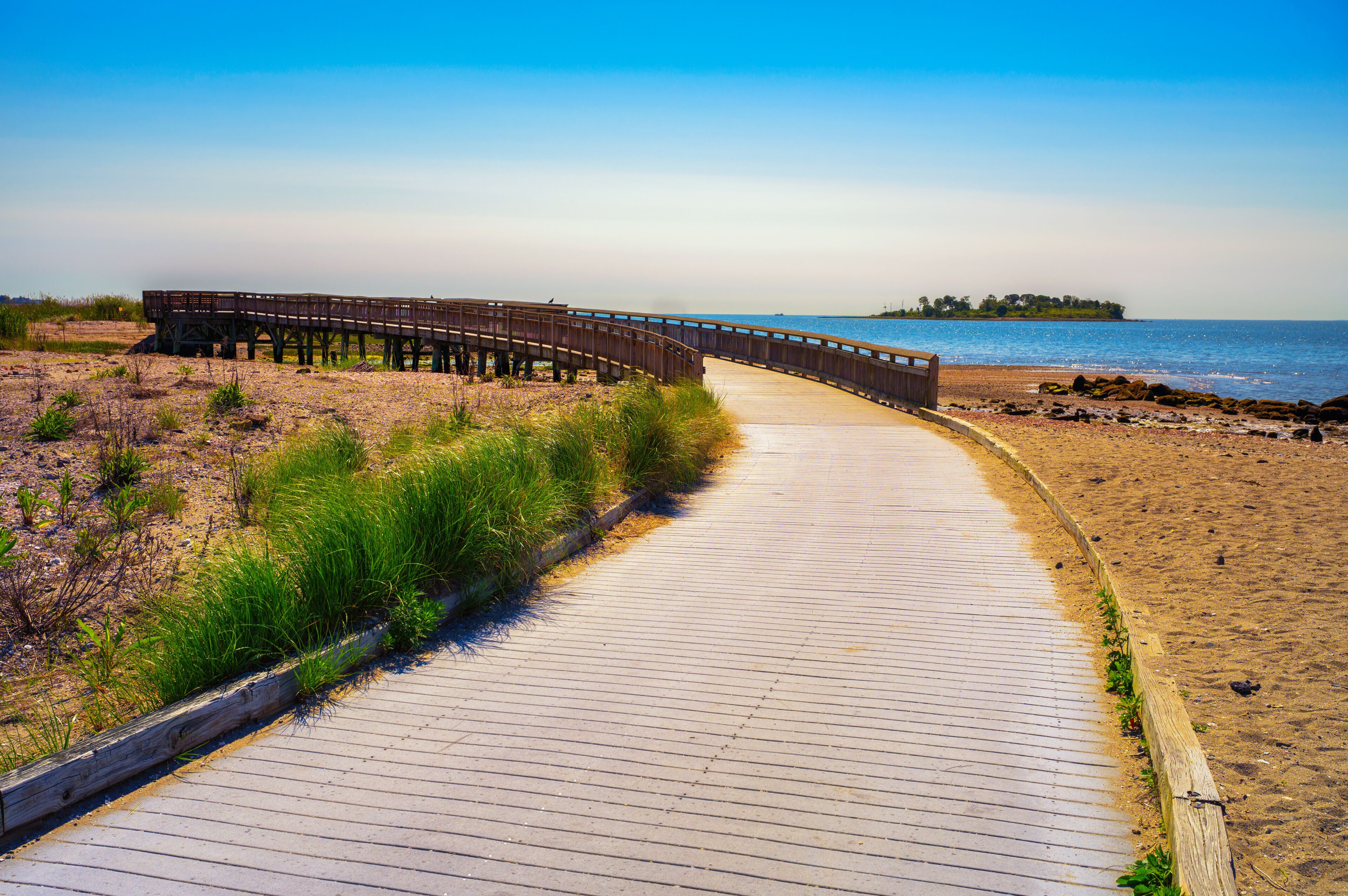 Curving wooden boardwalk along sandy beach at Silver Sands State Park in Milford, Connecticut, with calm blue water of Atlantic Ocean, coastal grass, and Charles Island visible in the background.