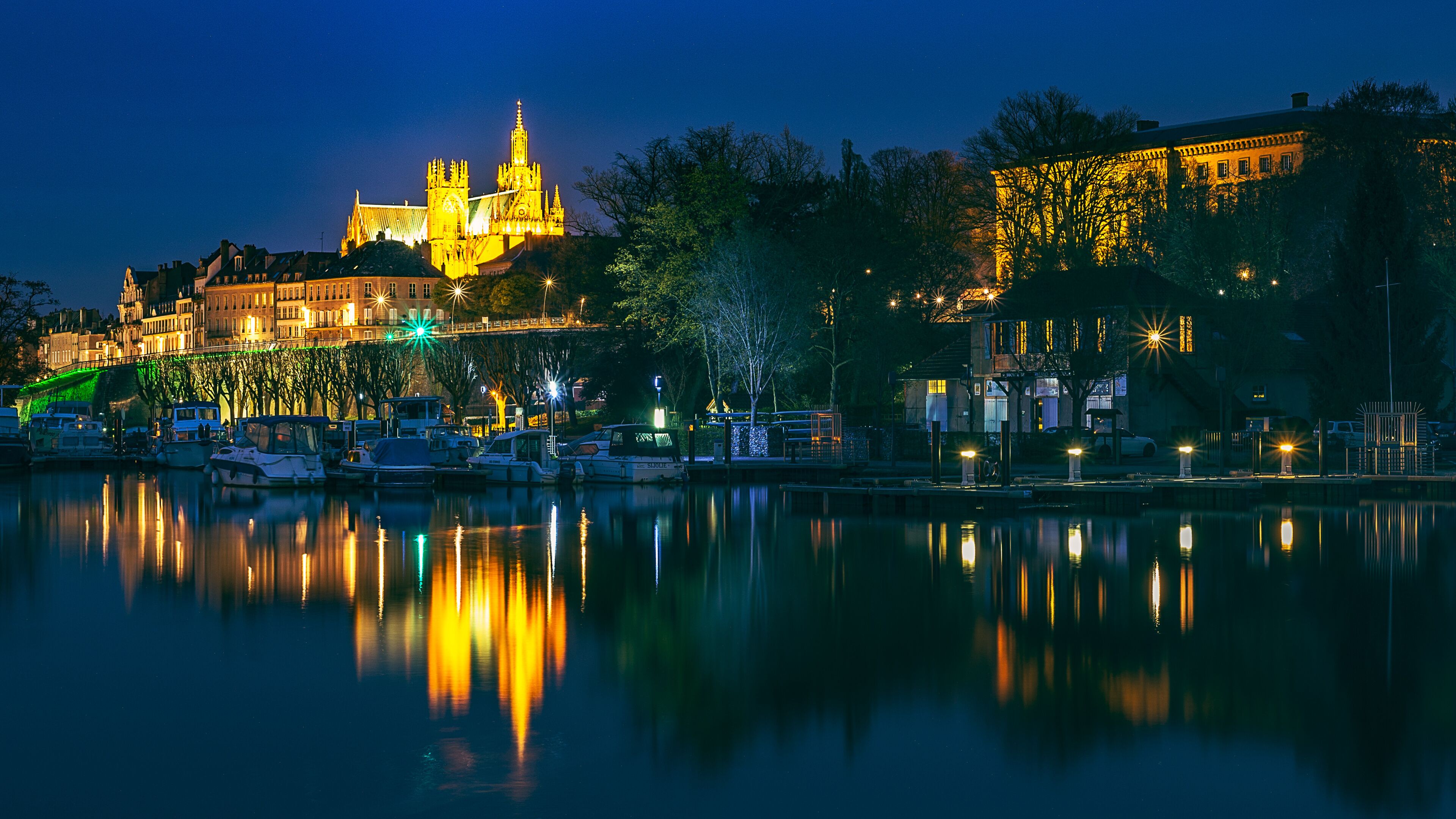 A nighttime cityscape with a prominent illuminated castle on a hill, a calm river reflecting the lights, and a bridge connecting the two sides.