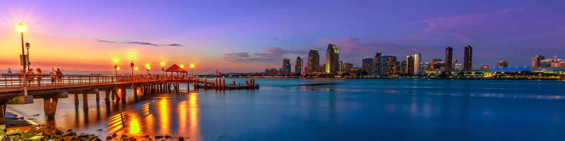 A city skyline at dusk with a bridge and pier reflecting city lights on the water.