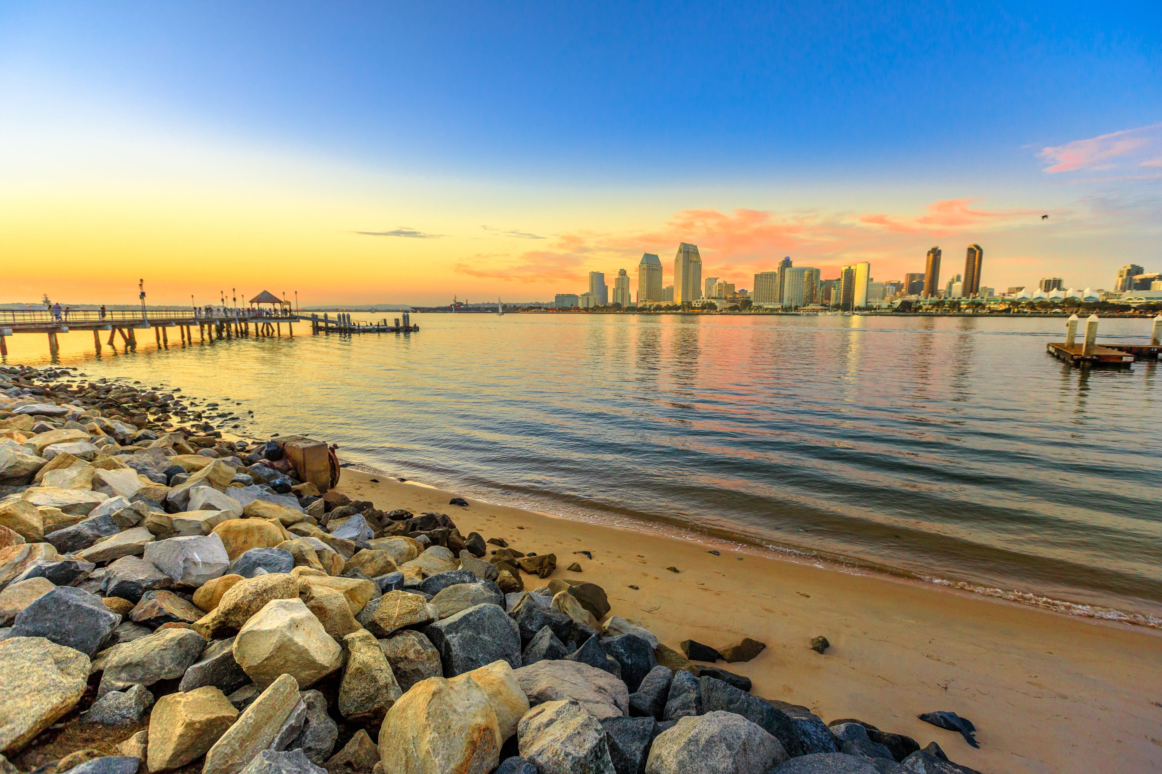 A rocky shoreline with a pier extending into calm waters, a city skyline in the background, and a sunset sky.