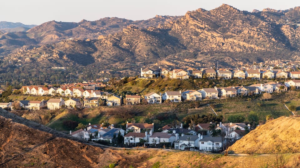 A hillside residential area with houses, a mountain range in the background, and a clear sky.