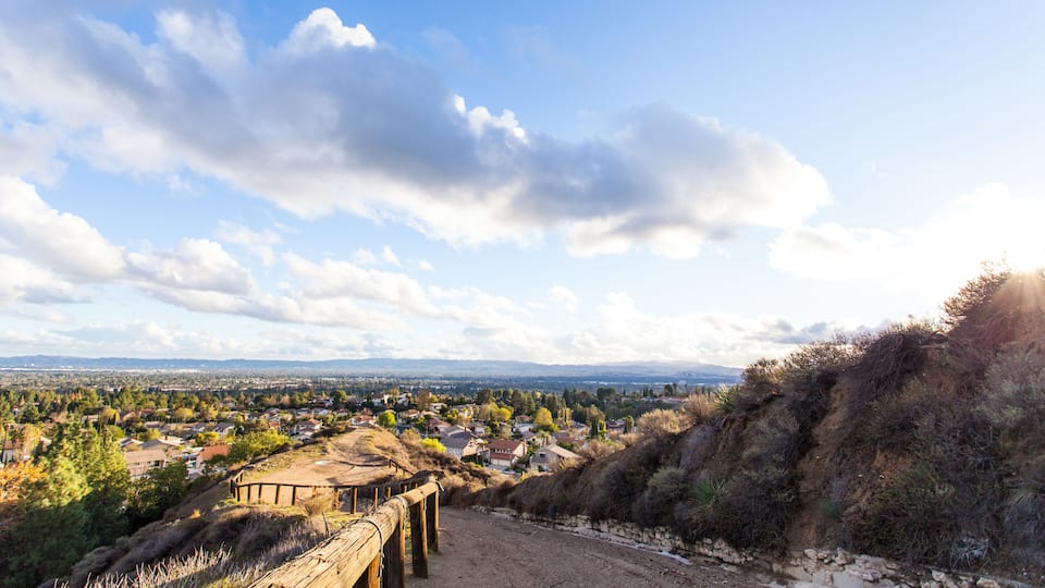 A rural landscape with a dirt road, wooden fence, and a distant view of houses and hills under a partly cloudy sky.