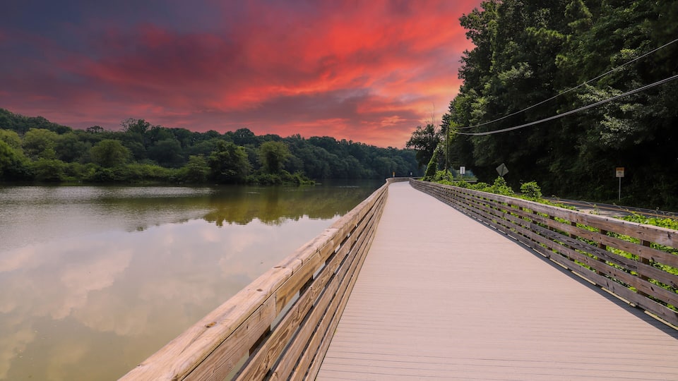 a long winding boardwalk along the Chattahoochee river with a wooden rail along the sides, still river water, lush green trees with powerful clouds at Roswell Riverwalk Boardwalk in Roswell Georgia