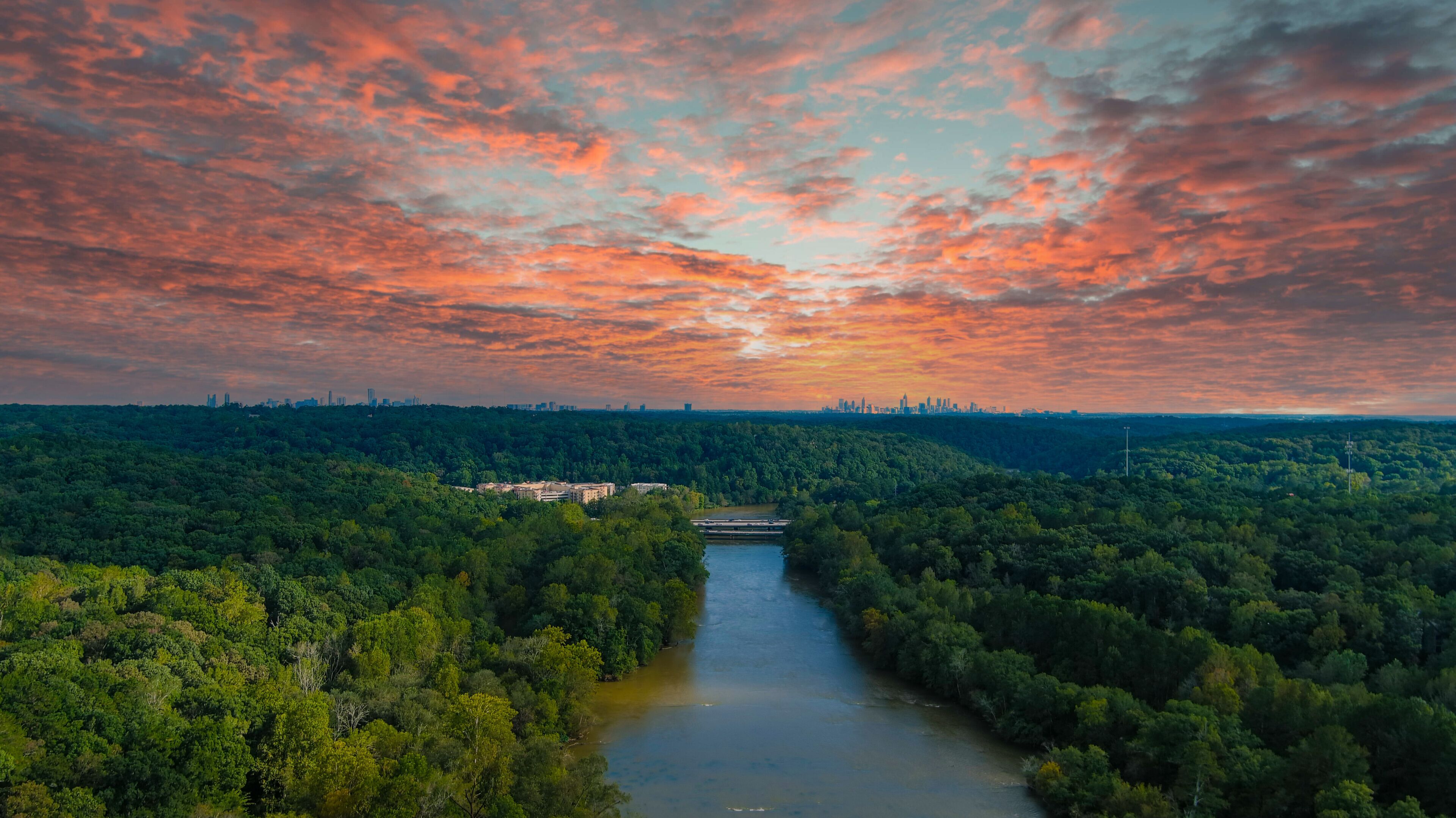 A river winding through a dense forest with a city skyline in the distance under a colorful sunset sky.