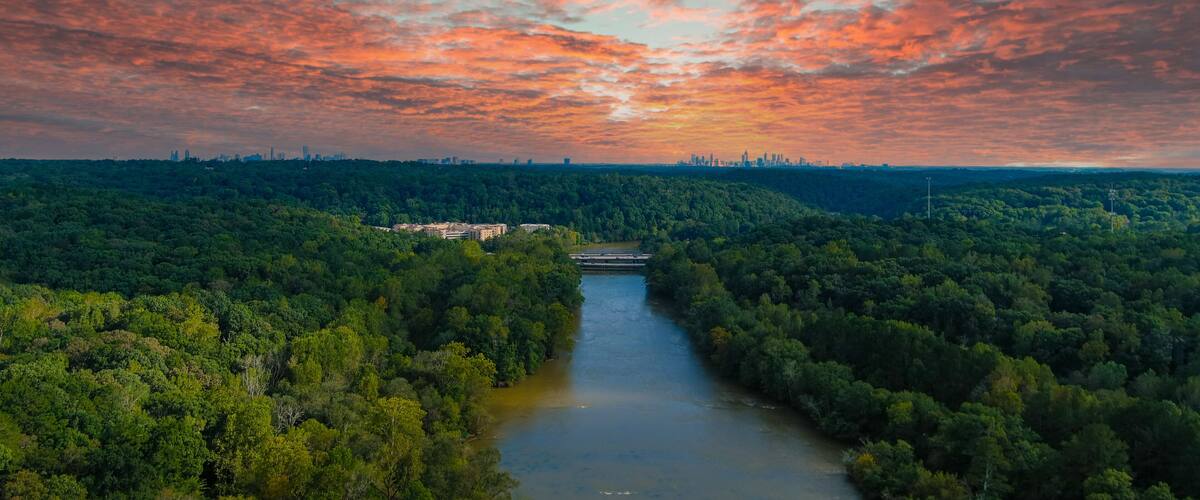 A river winding through a dense forest with a city skyline in the distance under a colorful sunset sky.