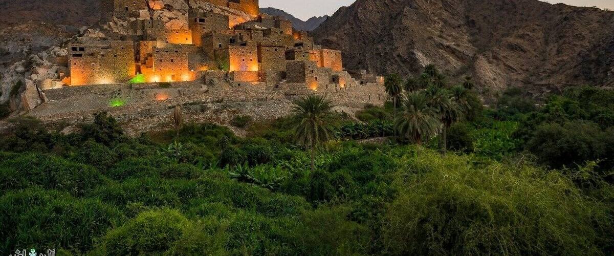 A historic stone fortress with illuminated windows, surrounded by lush greenery and rugged mountains.