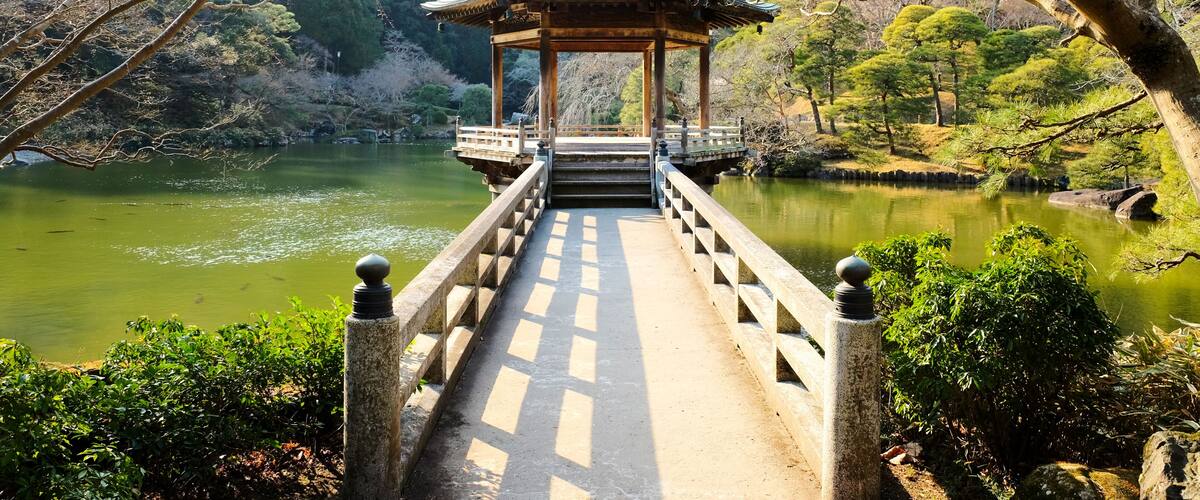 A traditional Japanese pavilion over a bridge leading to a calm lake surrounded by trees.